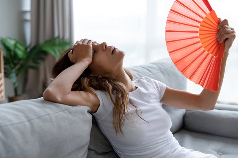 Why Is My AC Blowing Hot Air? Overheated young woman fanning herself with a paper fan in her living room on her couch.