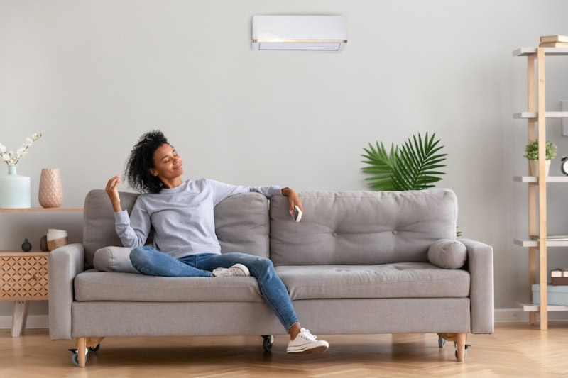 Ductless Mini Splits for Comfortable and Healthy Living. African American relaxed woman sitting on comfortable couch in living room at modern home holds air conditioner remote control enjoying breathing fresh cool air at summer or warm air at winter season.